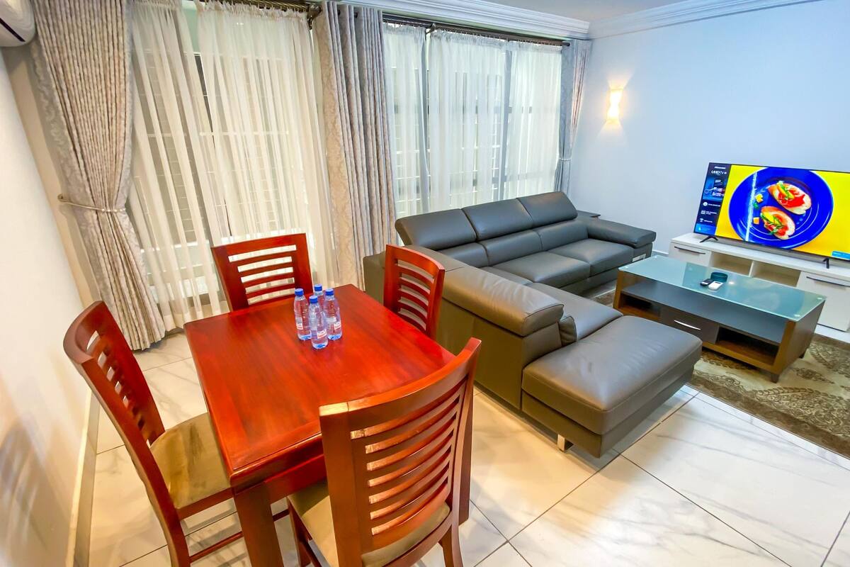 A living room with a sofa, a dining table with chairs, a TV, and a window with curtains.