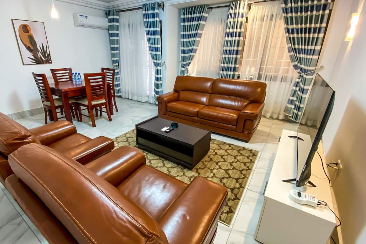 A living room with a brown leather sofa, a coffee table, a TV, and a dining area with a wooden table and chairs.