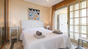 A hotel room with a large bed, two bedside tables with lamps, a window with curtains, and a butterfly wall art piece. - AirCon Apartment · Sea Views  · Pool · UHC CALA DORADA (Salou)
