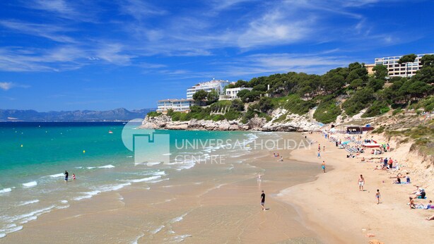 A beach with clear turquoise waters, people sunbathing, and buildings on a hillside. - AirCon Apartment · Sea Views  · Pool · UHC CALA DORADA (Salou)