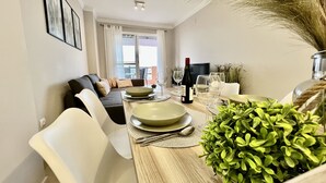 Uma área de refeições com uma mesa de madeira posta para dois, um sofá e uma TV ao fundo.