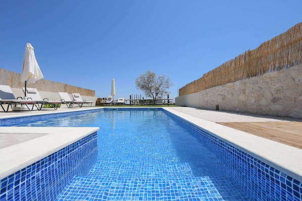 Une piscine entourée de murs en pierre et d’une terrasse en bois, avec des chaises longues et des parasols.