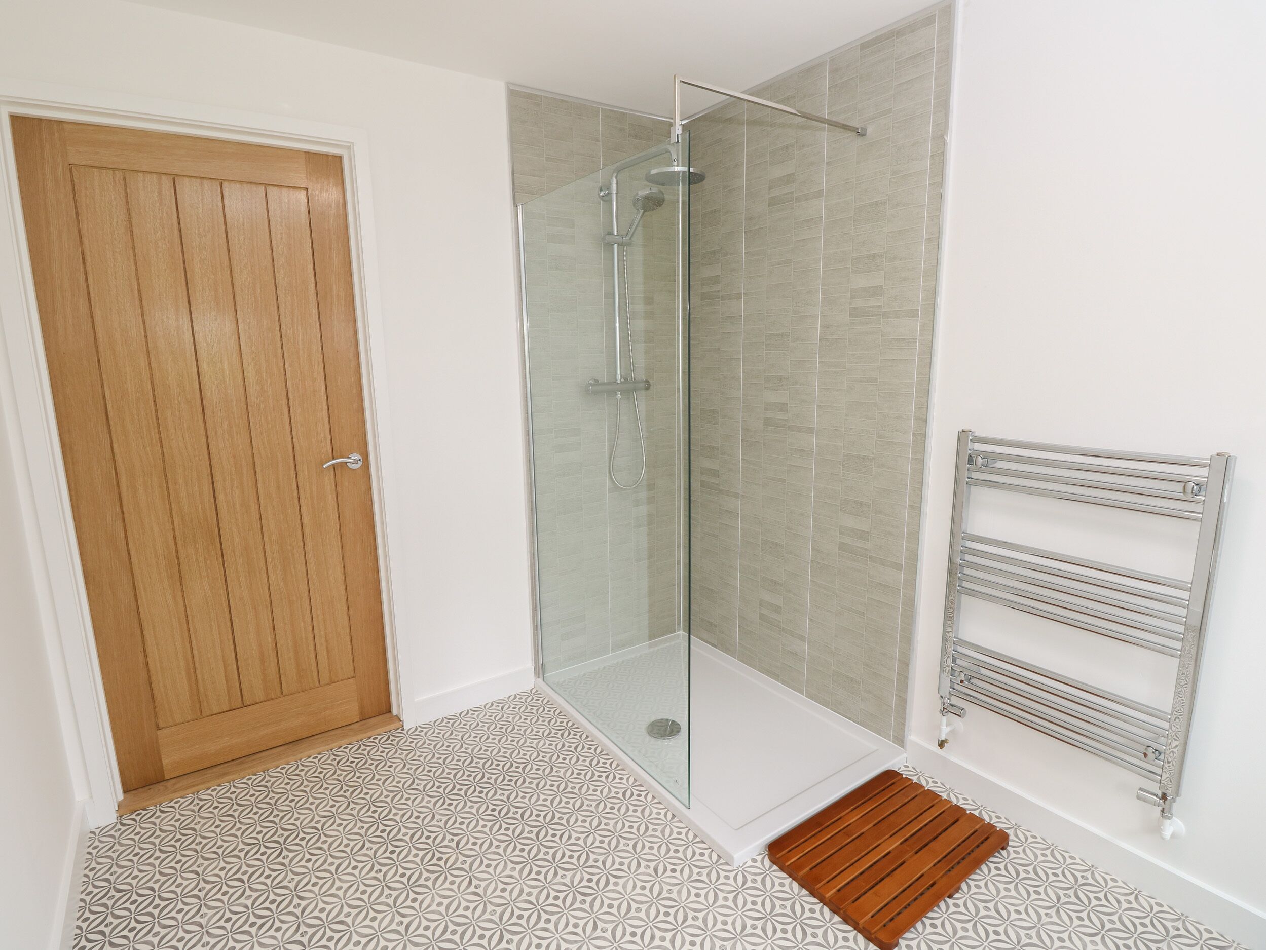 A bathroom with a glass shower enclosure, a wooden door, a patterned floor, and a metal towel rack.