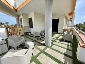 Et overdækket udendørs område med moderne, hvide møbler, herunder stole og en bænk, placeret på en gulv med et mønster af grønne og beige fliser.
