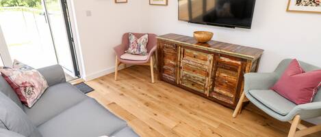 Una sala de estar moderna con un sofá gris, un sillón rosa y un sillón gris con un cojín rojo. Un mueble de madera con cajones y un televisor montado en la pared.