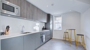 Een moderne keuken met grijze kastjes, een ingebouwde magnetron, een spoelbak en een raam. Drie gouden krukken staan bij het raam.