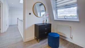 Een moderne slaapkamer met een groot raam, een zwarte dressoir, een ronde spiegel en een blauwe voetenbank.