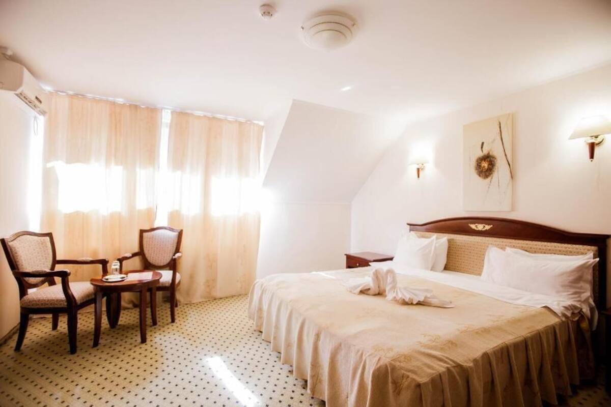 A hotel room with a bed, two chairs, a small table, a nightstand, and a wall-mounted lamp.