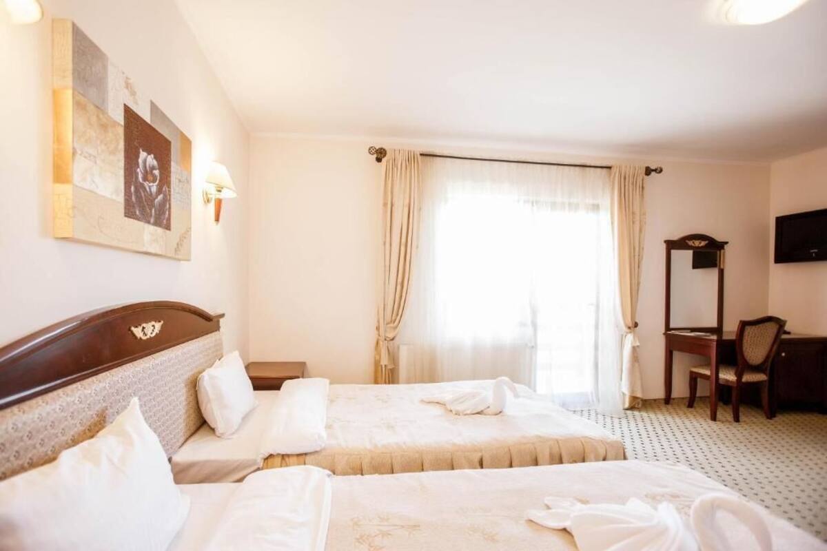 A hotel room with two beds, a desk, a chair, a TV, and a large window with curtains.