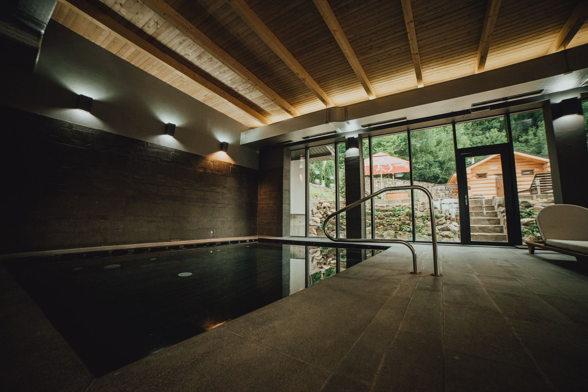 An indoor swimming pool with a wooden ceiling and stone walls, a glass door leading outside, and a chair near the pool.