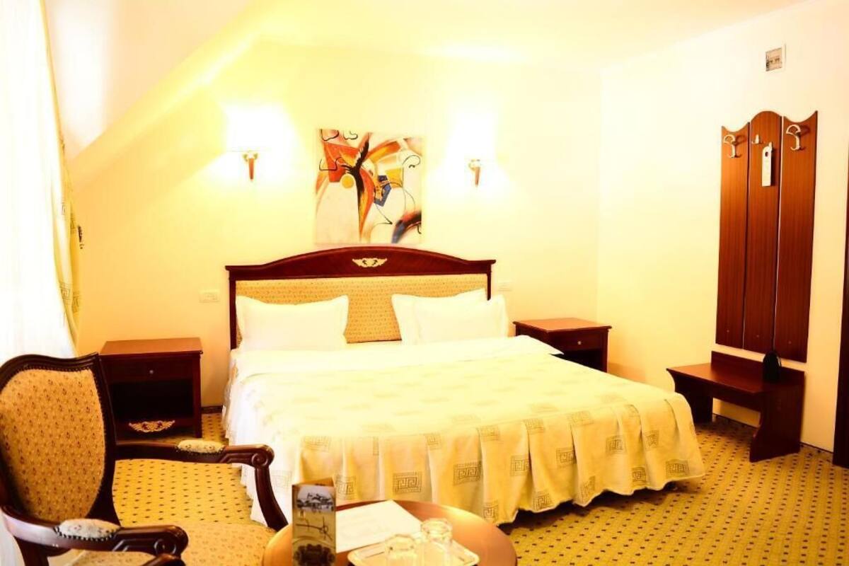 A hotel room with a large bed, a wooden nightstand, a chair, a desk, and a wardrobe.