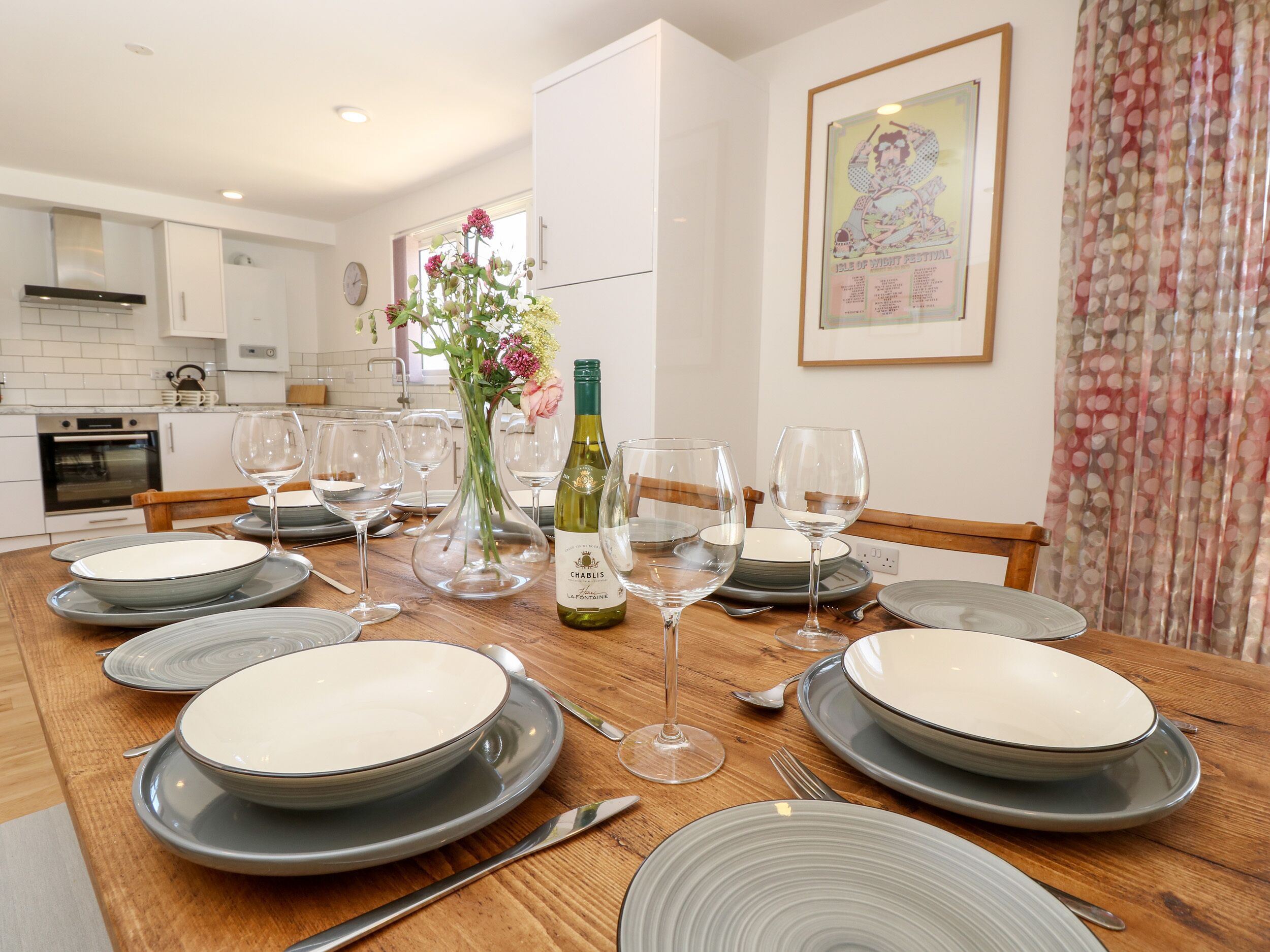 A dining table set with plates, glasses, and a bottle of wine, with a kitchen in the background.