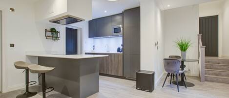 A modern kitchen with a central island, bar stools, a dining table with chairs, and a staircase leading to another level.