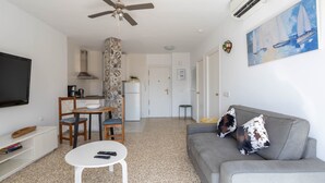 A living room with a grey sofa, a television, a ceiling fan, and a kitchen area in the background.