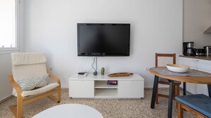 A modern, minimalist room with a flat-screen TV mounted on the wall, a wooden chair with a cushion, a small white cabinet, and a round table with a bowl and a rolling pin.
