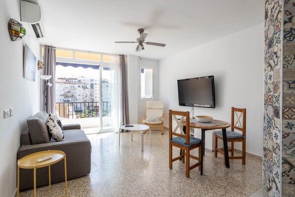 A modern living room with a sofa, a TV, a ceiling fan, and a balcony with a view of the city.