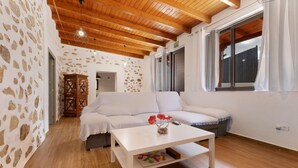 Habitació amb paret de pedra, bigues de fusta al sostre, un sofà blanc, una taula blanca i una finestra amb cortines.