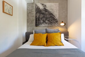 Un lit recouvert d’une couvre-lit blanche et grise, deux coussins moutarde, un coussin gris et une couverture grise. Au-dessus du lit, un tableau abstrait en noir et blanc est accroché au mur, et une petite table avec une lampe se trouve à côté du lit.