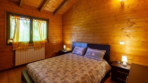 A wooden cabin bedroom with a bed, nightstands, and a window with curtains. - Chalet 'Mirtillo' – Villaggio Anemone with Mountain Views and Private Terrace (Capanne)