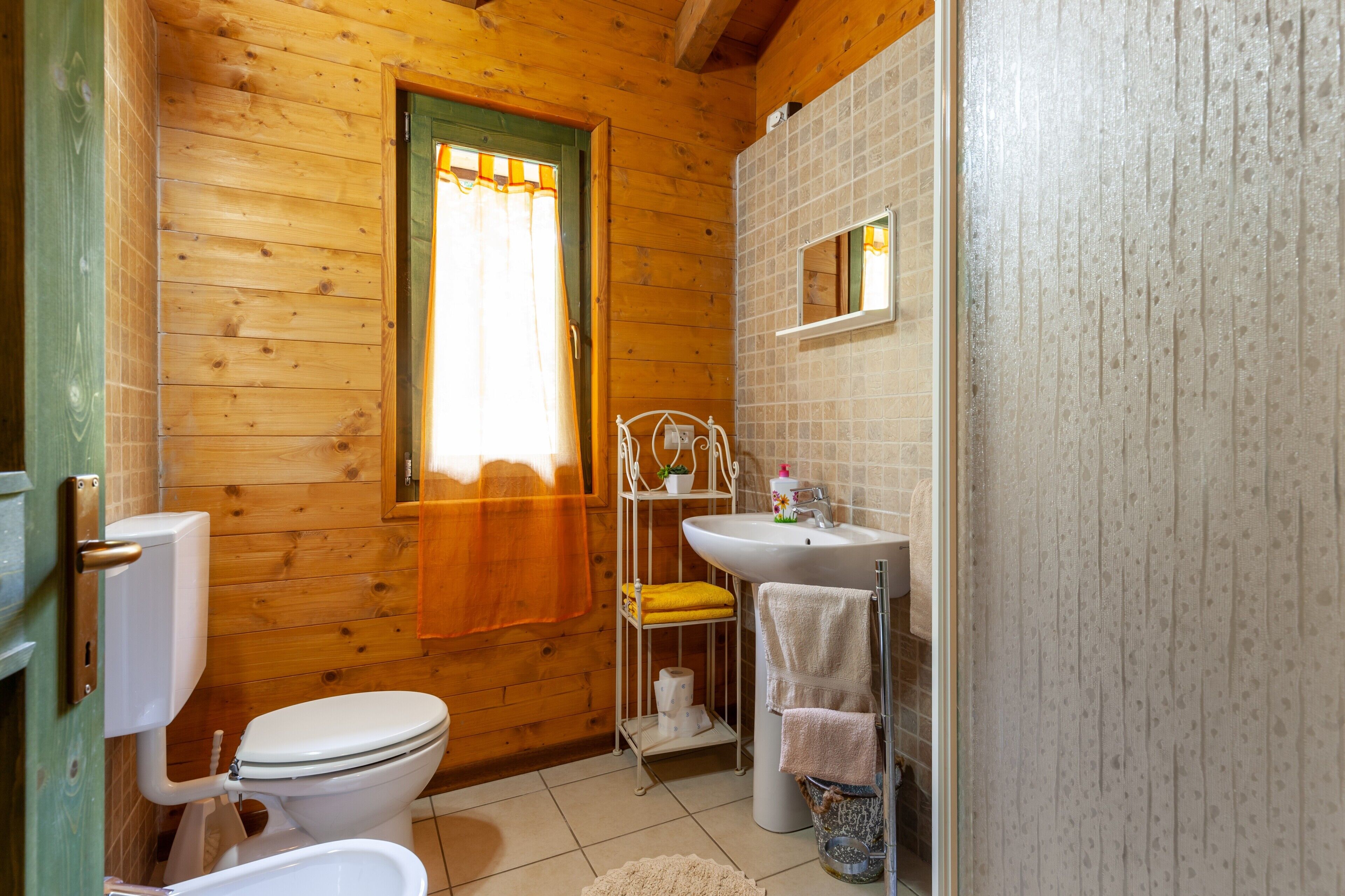 A wooden cabin bathroom with a toilet, sink, and window with curtains.