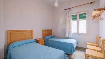 Un dormitorio con dos camas, una mesita de noche de madera, una ventana con cortinas y una silla de madera.