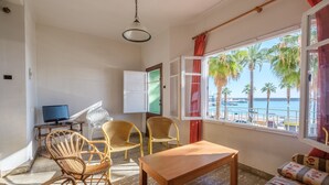 Ein Zimmer mit Holztisch, Korbstühlen, einer Couch, einem Fernseher und einem Fenster mit Blick auf Palmen und das Meer.
