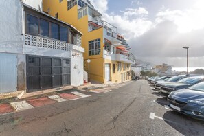 Biler parkeret langs en gade med høje bygninger, altaner og et hvidt hegn.