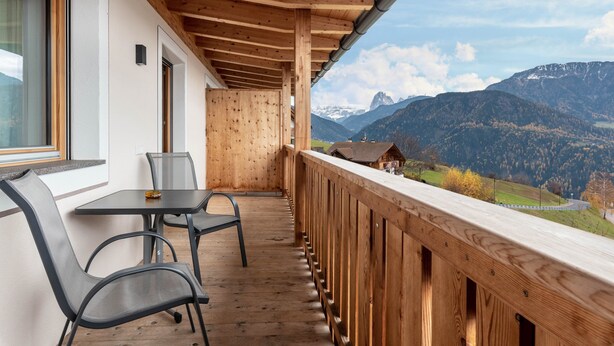 A balcony with a wooden railing, a table, and two chairs, overlooking a mountainous landscape.