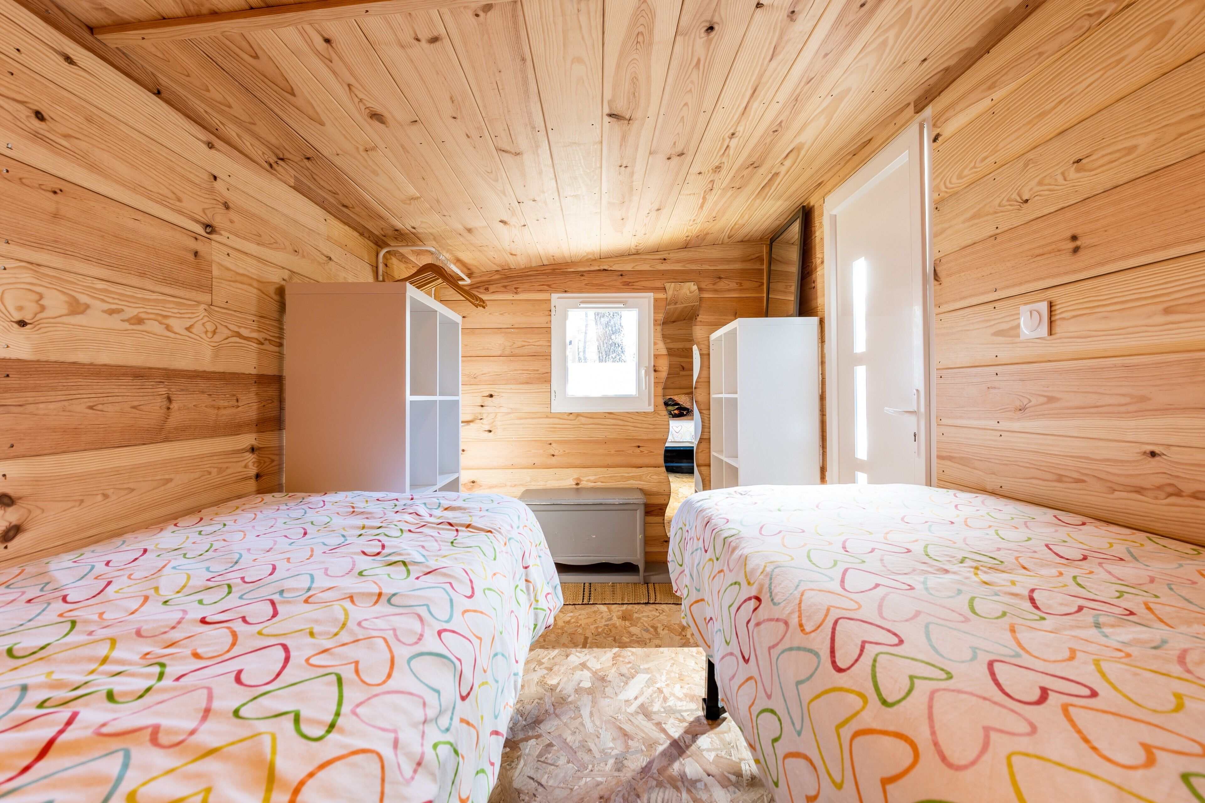 Una cabaña con paredes de madera, dos camas con ropa de cama de estampado de corazones coloridos, un ventanal pequeño y un calentador blanco.