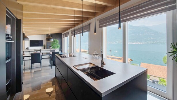 A modern kitchen with a large island, wooden ceiling beams, and a view of the water.