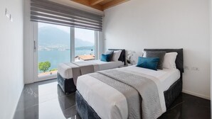 A bedroom with two beds, a large window with a view of the sea and mountains, and a wooden ceiling.