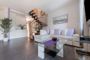 A modern living room with a glass coffee table, white sofa, and a staircase with wooden railings. - Luxury Lakefront Maisonette with Lake View & Wi-Fi (Lezzeno)