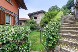 A stone building with a red roof, a grassy yard, and a staircase leading up. - Luxury Lakefront Maisonette with Lake View & Wi-Fi (Lezzeno)