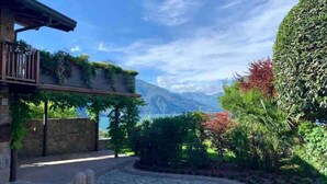 A stone pathway leading to a building with a balcony, surrounded by lush greenery and a mountain view.