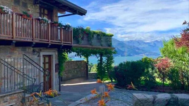 A two-story building with a balcony, stone walls, and a view of a lake and mountains.