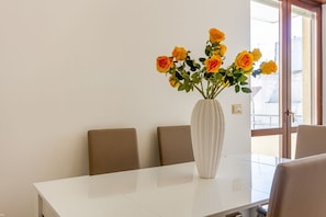 A dining table with a white vase containing yellow roses, brown chairs, and a window with a view of buildings.