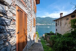 Un bâtiment en pierre avec des volets en bois, un escalier en pierre et une vue sur une étendue d’eau et des montagnes.