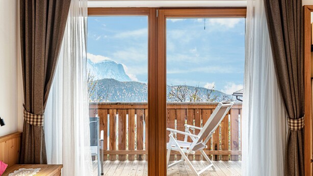 Ein Zimmer mit einem Bett, einem Nachttisch aus Holz, einem Fenster mit Vorhängen und einem Balkon mit einem Stuhl.