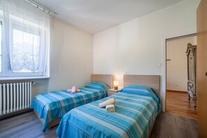Ein Schlafzimmer mit zwei Betten, einem Fenster mit Vorhängen, einem Heizkörper, einem Nachttisch mit Lampe und Holzboden.