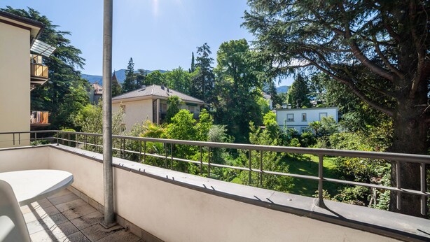 A balcony with a metal railing, a white table, and a view of a residential area with trees and houses. - Residence Hubertus Apartment 2 with Balcony & Wi-Fi (Meran)