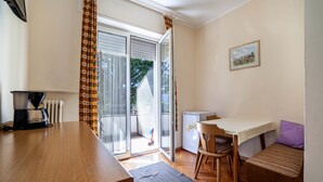 A small room with a wooden table, a chair, a bench, a radiator, a coffee maker, and a window with curtains. - Residence Hubertus Apartment 2 with Balcony & Wi-Fi (Meran)