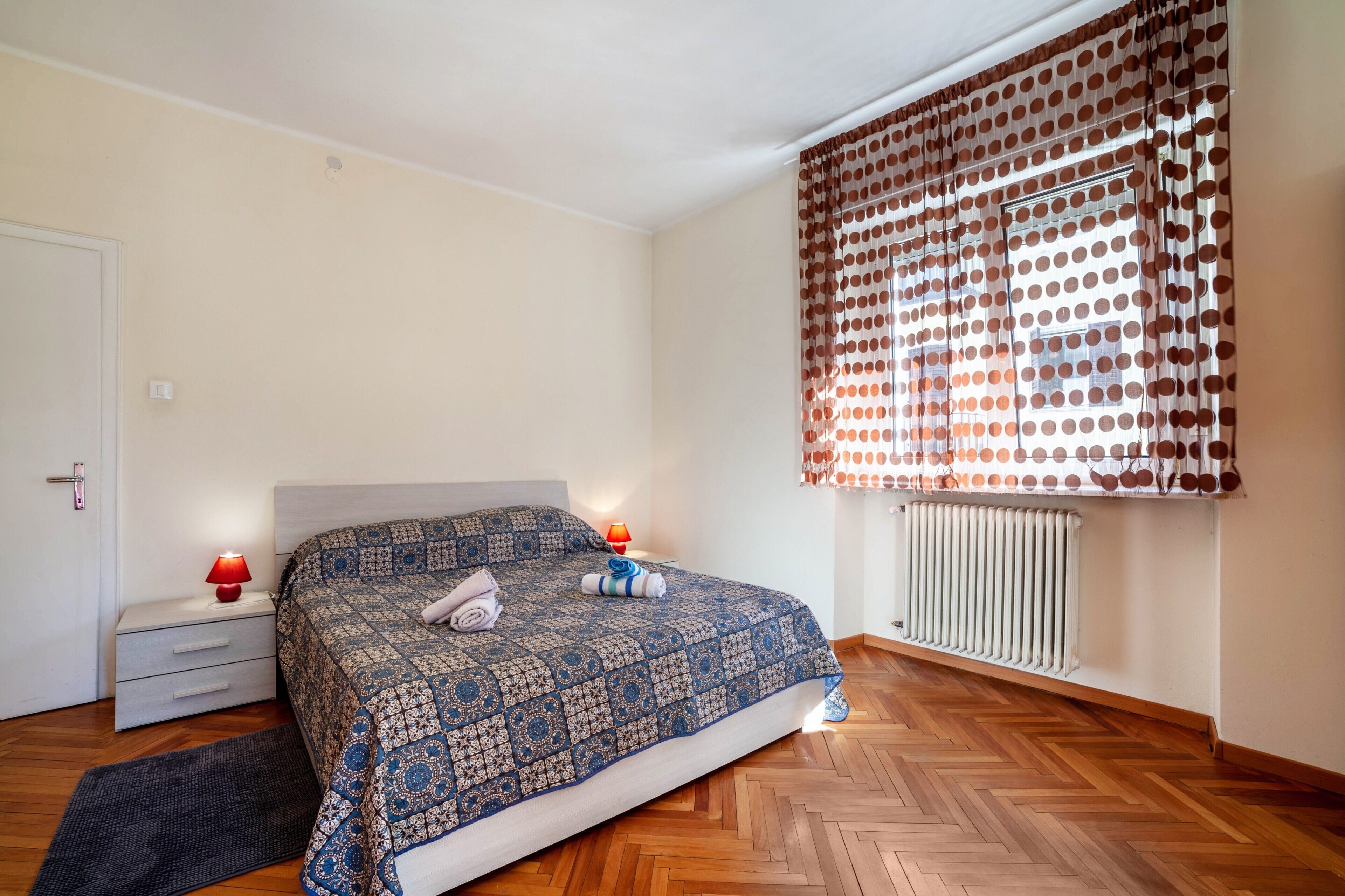 Ein Schlafzimmer mit einem gemusterten Bettüberwurf, einem Heizkörper, einem Fenster mit Punktevorhängen, einer Nachttischlampe und einem Holzboden.