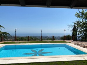 Une piscine rectangulaire aux motifs de mosaïques bleues, entourée d’un mur de briques et d’une clôture métallique, offrant une vue panoramique sur l’océan.