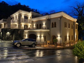 Une grande maison à étages, dotée d’une façade en pierre, d’un balcon et d’une voiture garée devant.