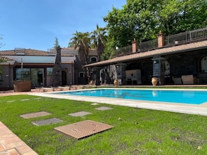 Una piscina con un patio de piedra, un área de estar al aire libre cubierta y un edificio con techo de tejas.