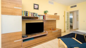 Een moderne woonkamer met een flatscreen televisie, een houten entertainmentkast, boeken, een kleine plant en een blauwe bank.