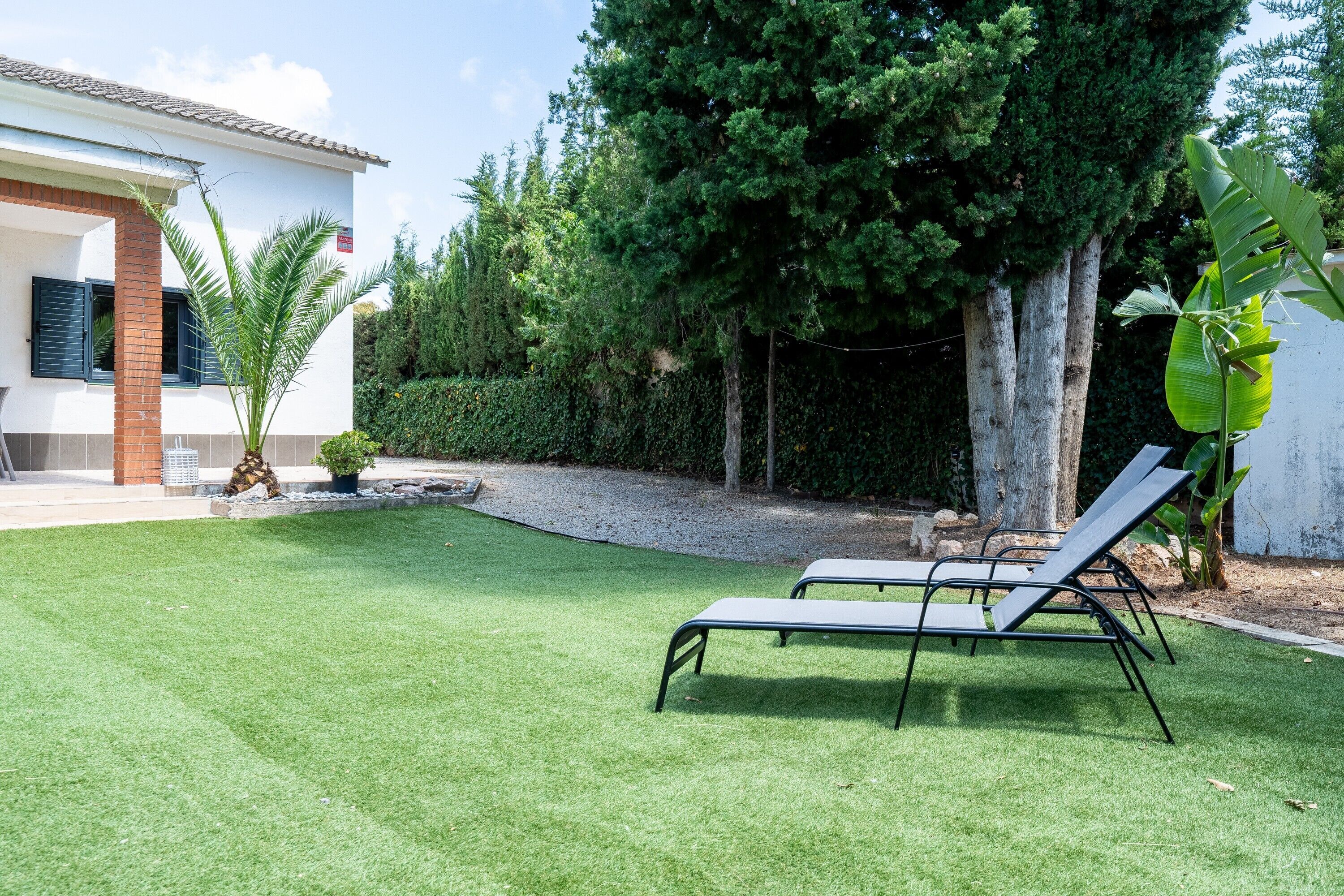 A well-maintained lawn with two lounge chairs, a white building with a tiled roof, and a dense green hedge.