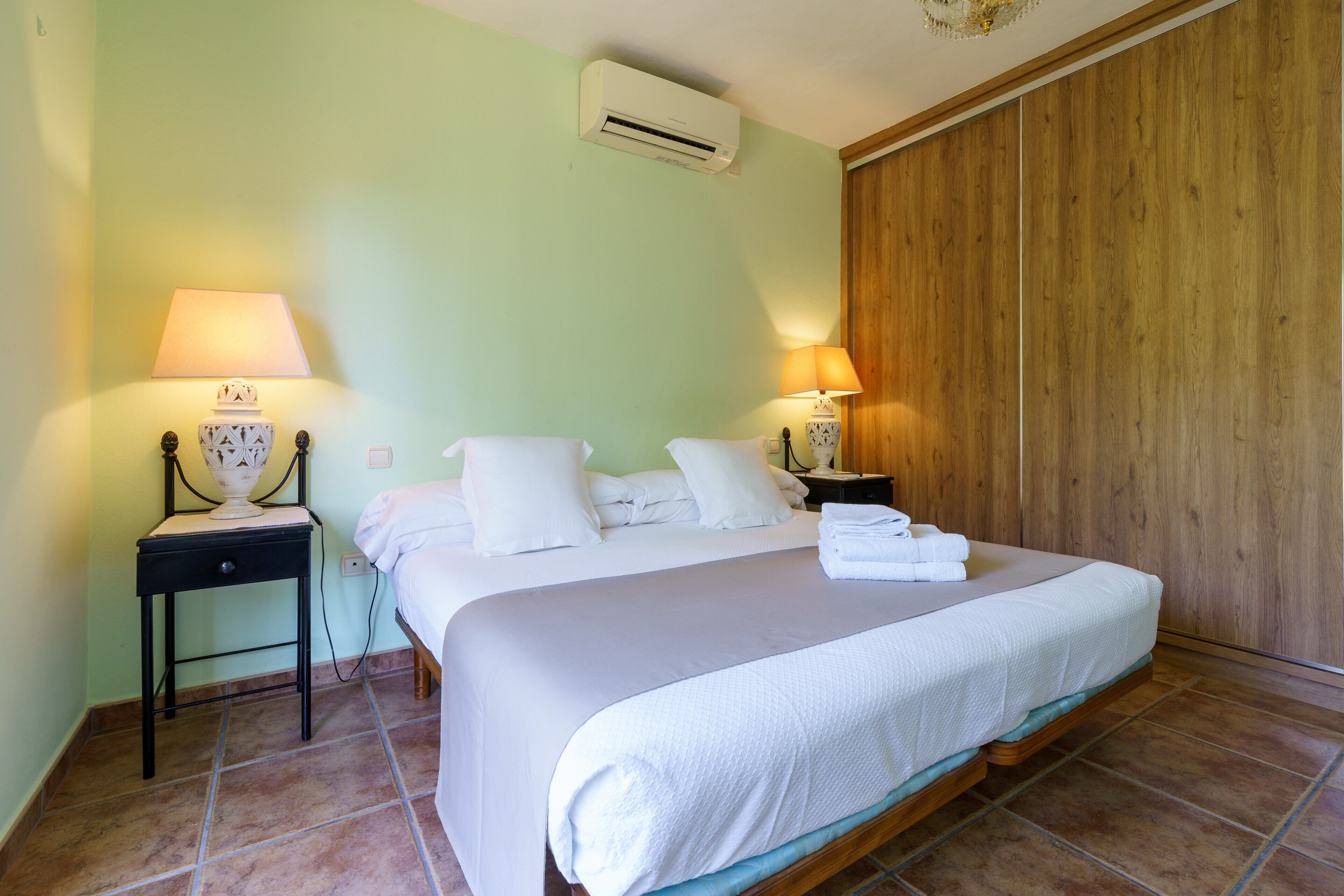 A hotel room with a bed, two bedside tables with lamps, a wall-mounted air conditioner, and a wooden panel wall.