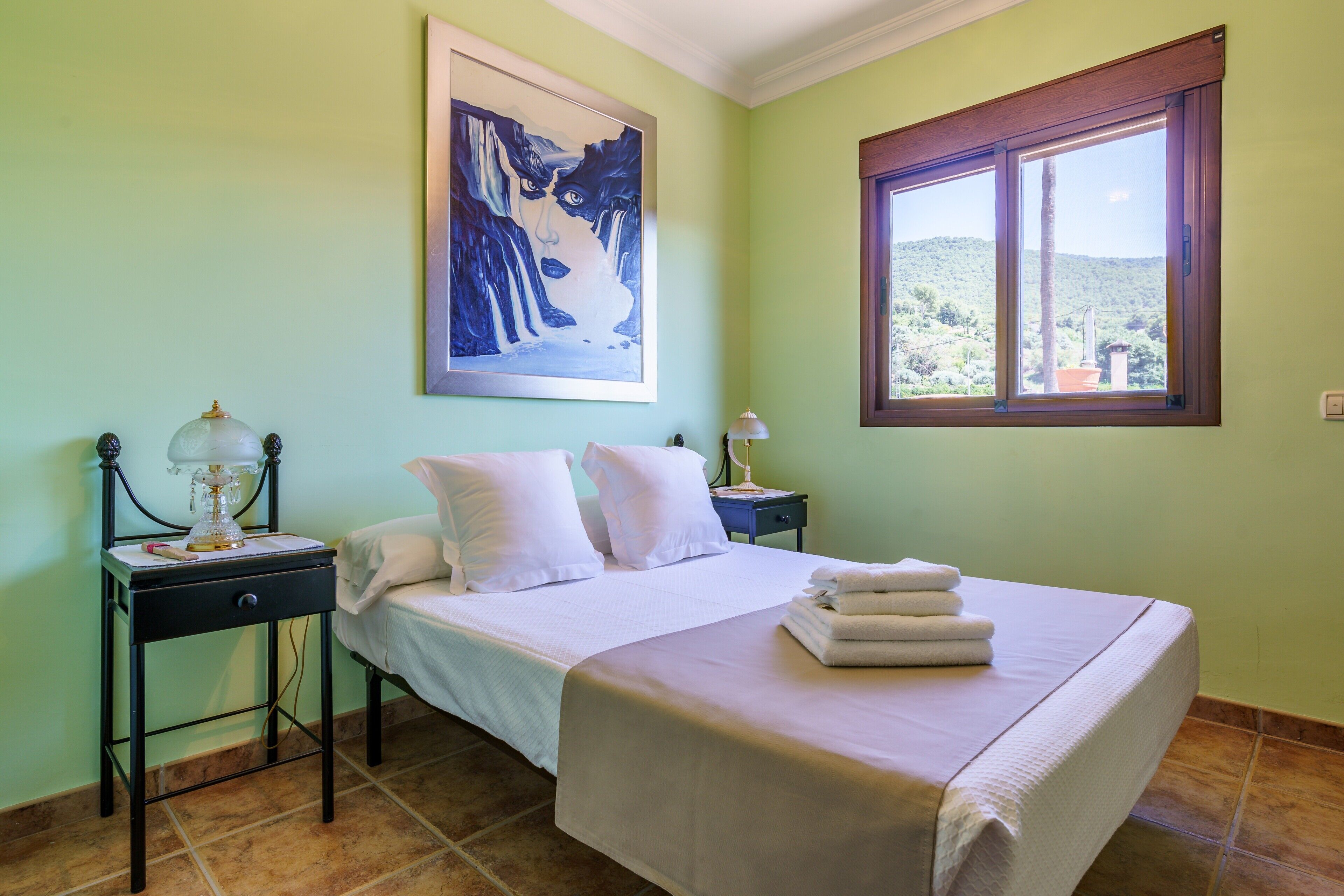 Un dormitorio con cama, mesita de noche, lámpara, un cuadro y ventana con vistas.