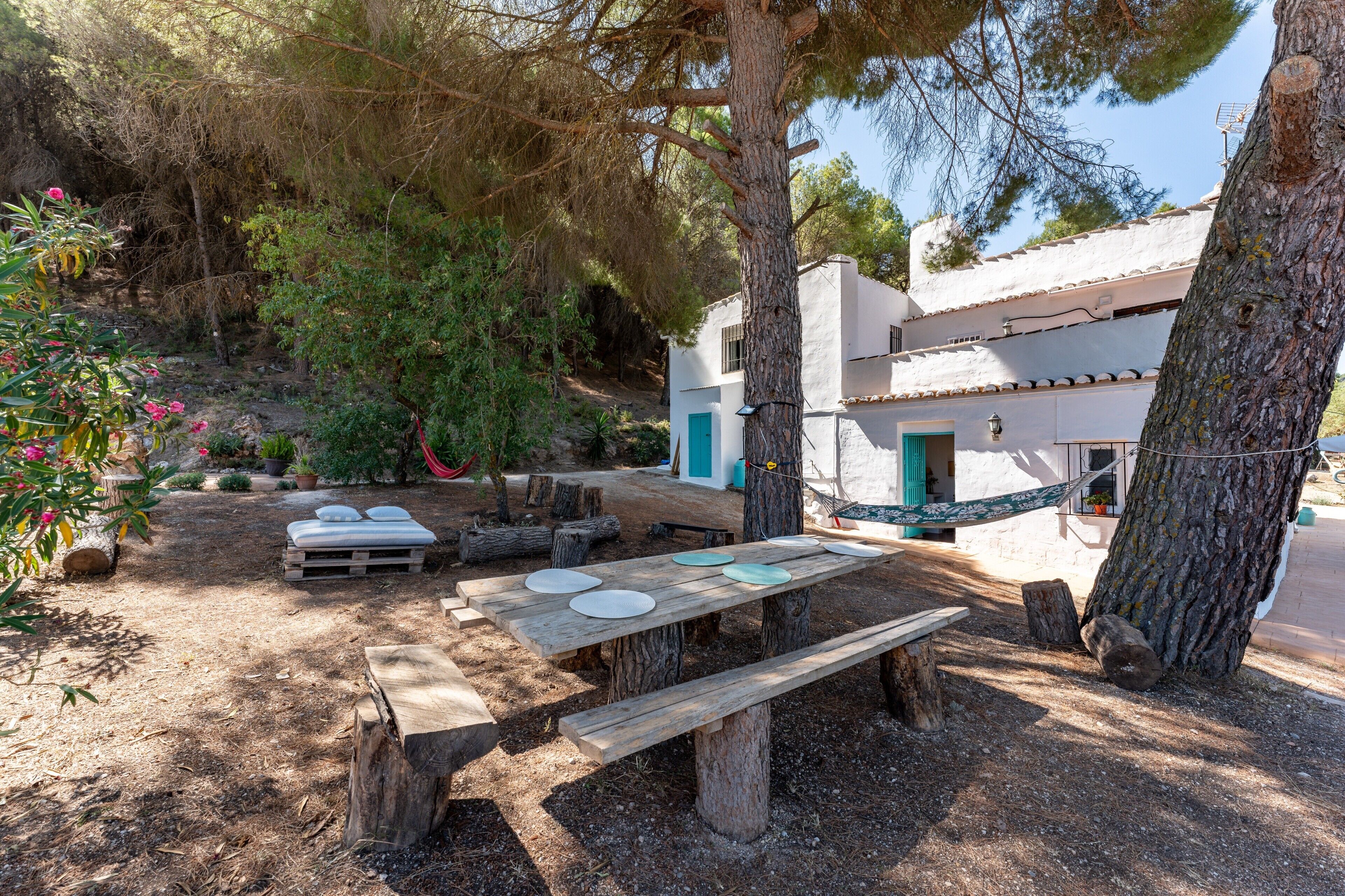 A rustic outdoor dining area with wooden benches and picnic tables, surrounded by trees and a white building with a blue door.